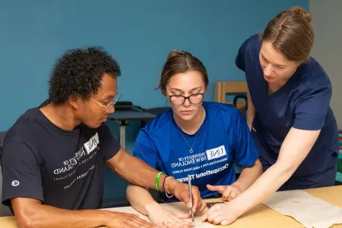 An O T student draws around a fellow student's wrist onto paper as a professor describes the process