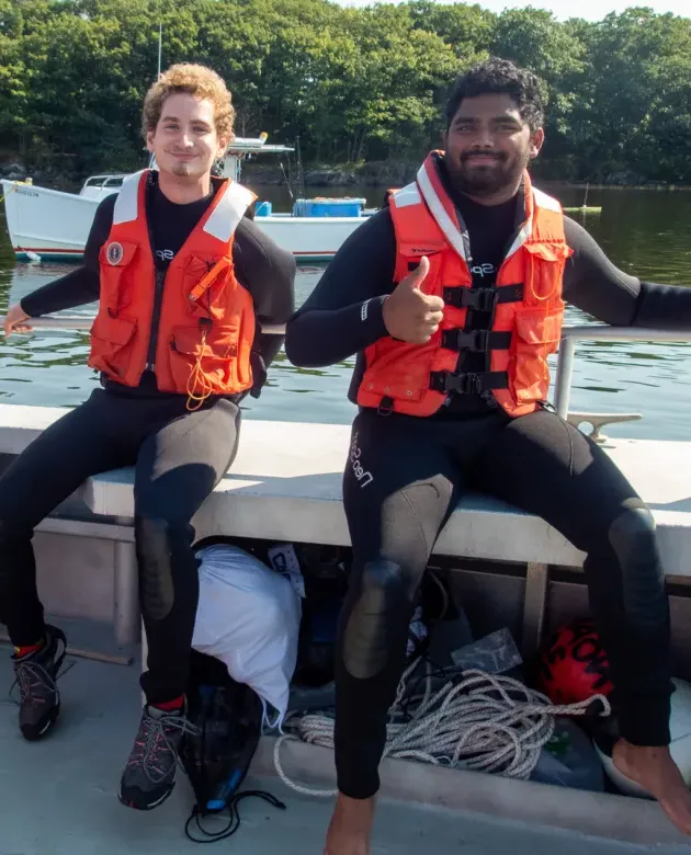 Two marine science students wearing wetsuits and orange life vests sit on the edge of a boat